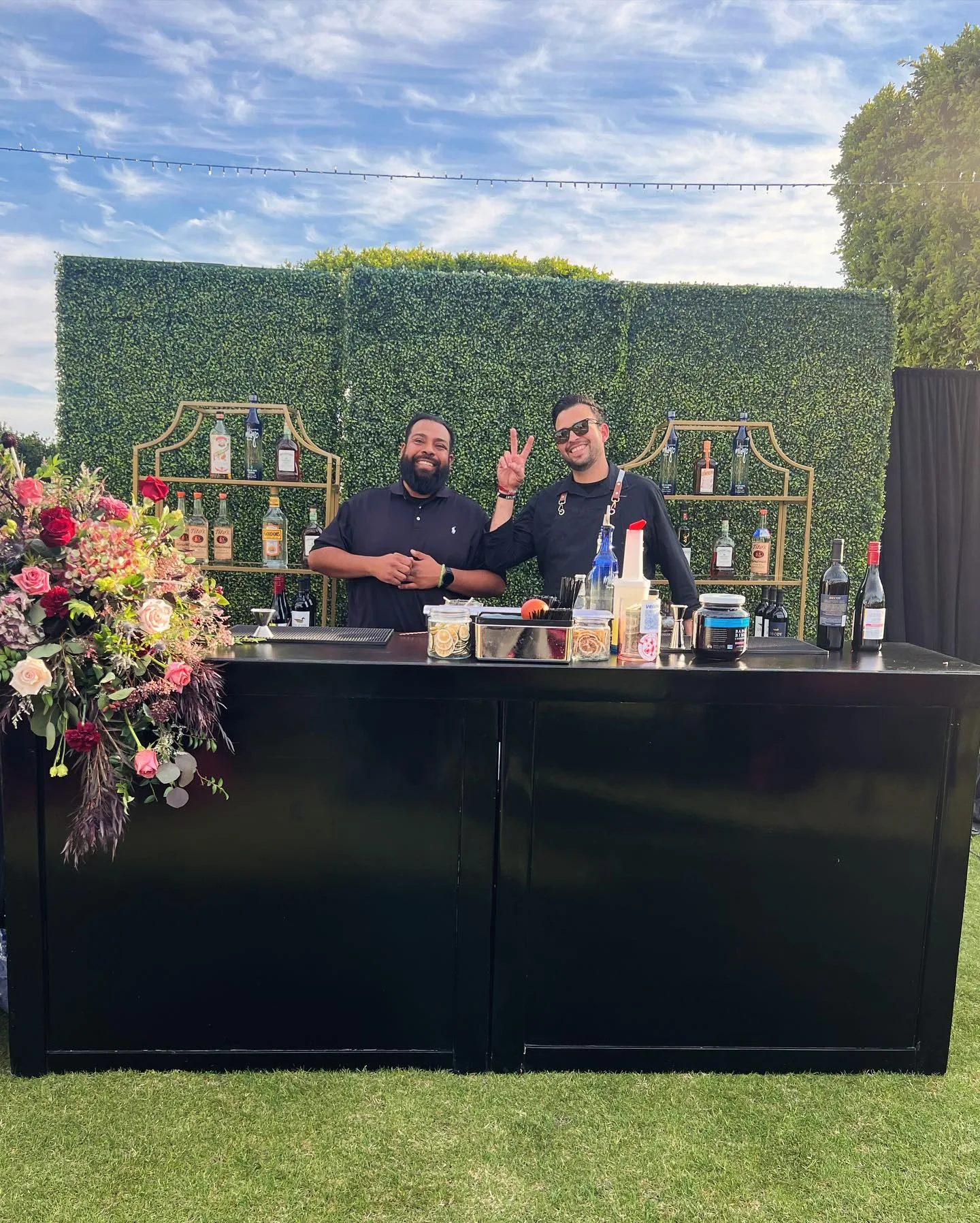 Two bartenders behind a black outdoor bar framed with colorful flowers and desert mountains