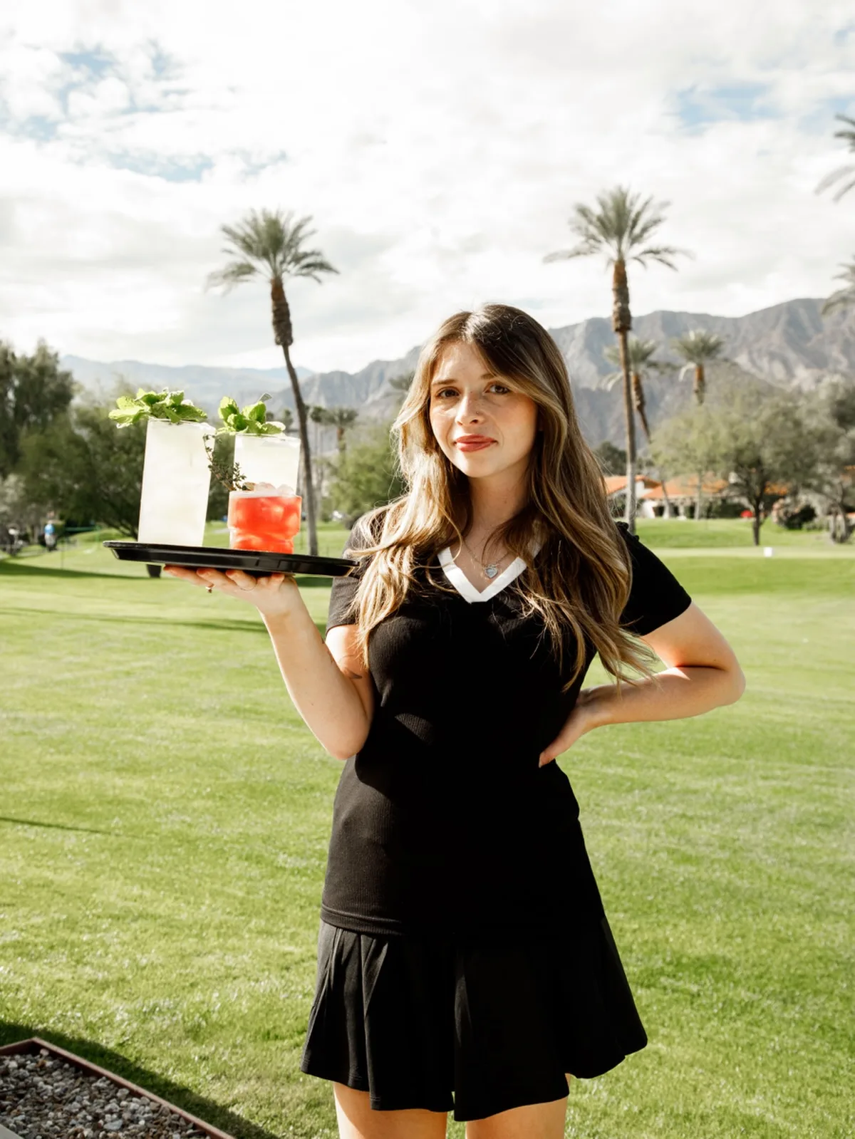 Event staff member carrying two colorful cocktails across a lawn with desert mountains behind her