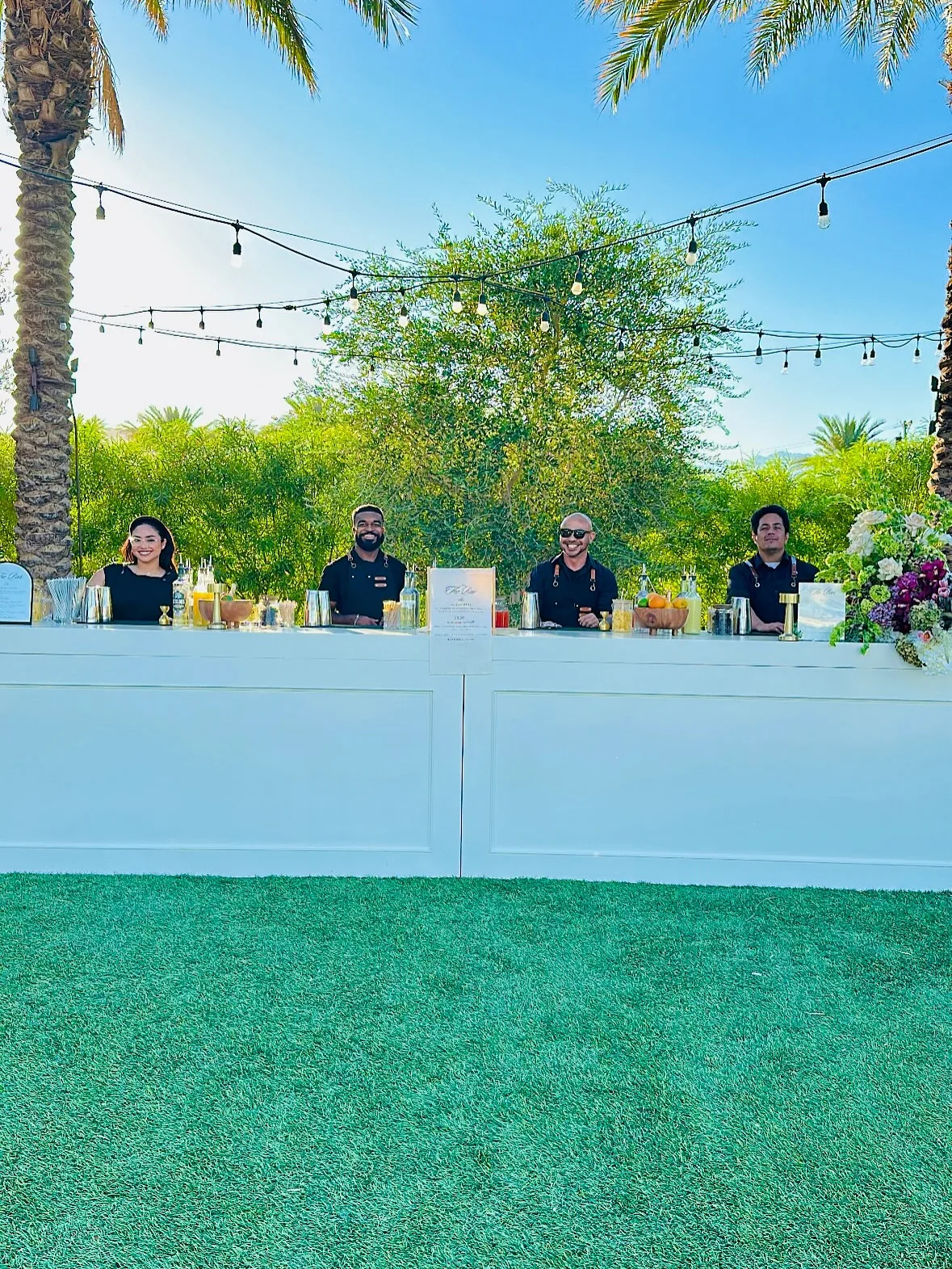 Four That’s The Spirit bartenders smiling behind a long outdoor bar beneath palm trees and string lights