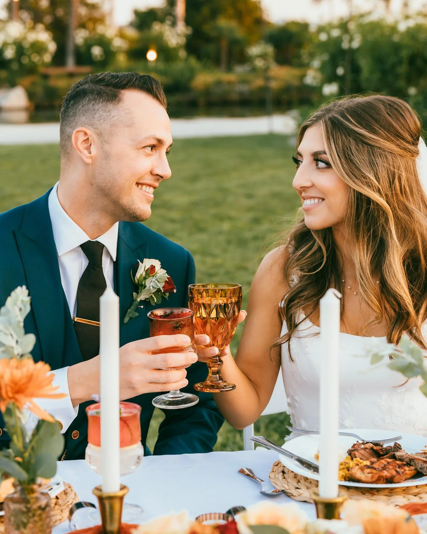Wedding couple smiling at one another while holding cocktails at their outdoor reception table