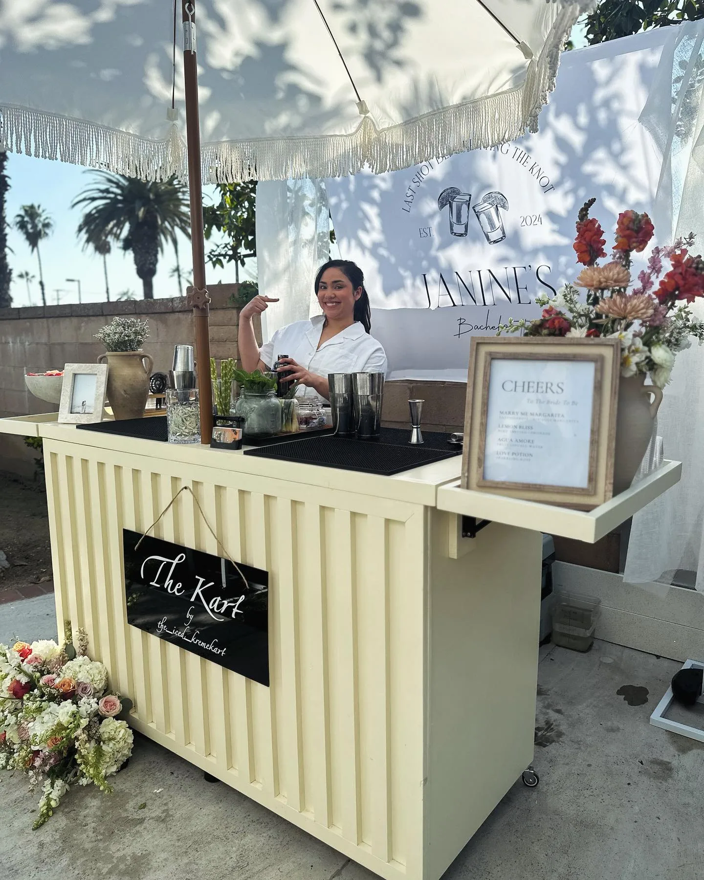 Cream mobile bar styled with flowers, cocktail ingredients, and a printed menu at an outdoor event