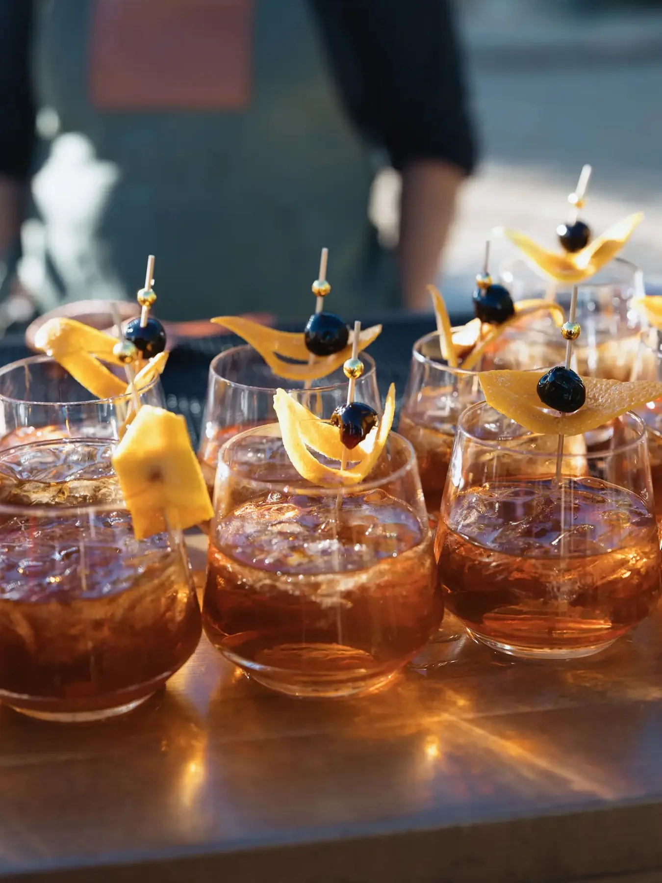 A sunlit row of amber cocktails garnished with orange peel and dark berries