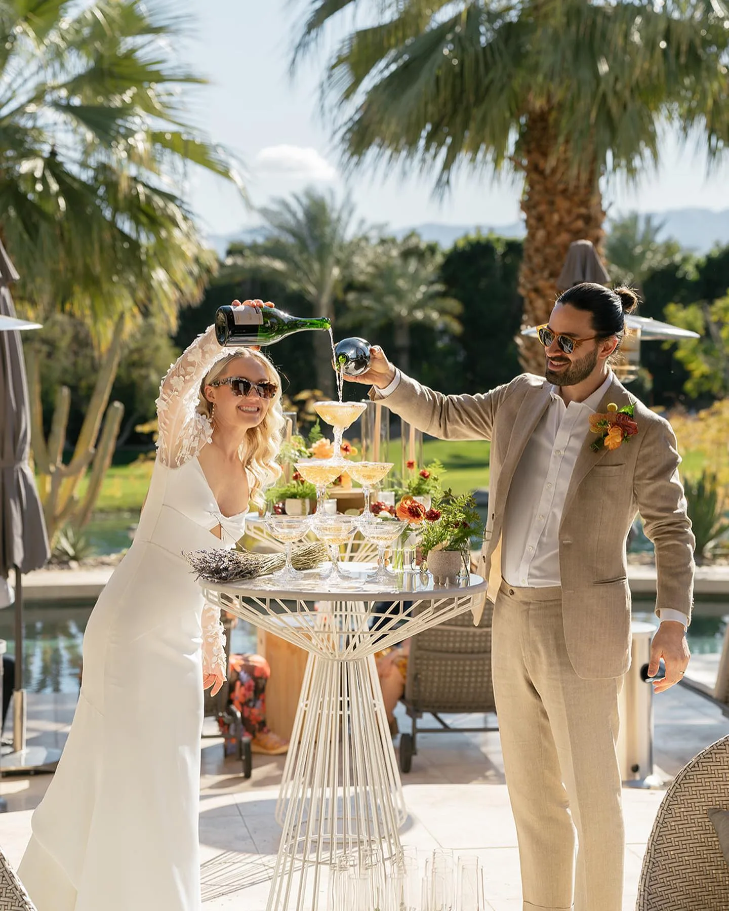 Newlyweds raising cocktails beside a flower-covered cocktail cart beneath Palm Springs palm trees