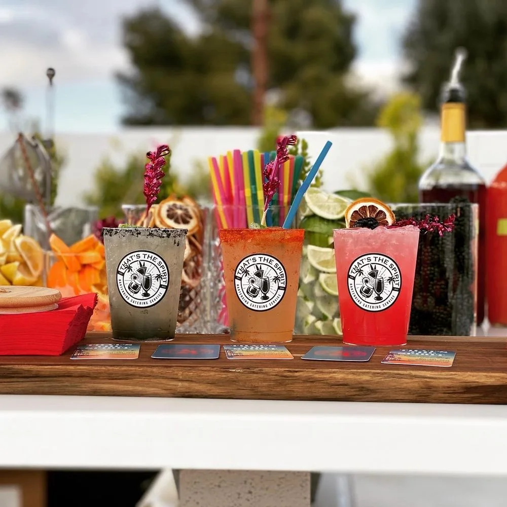 Three colorful cocktails with custom labels lined up on a wooden serving board