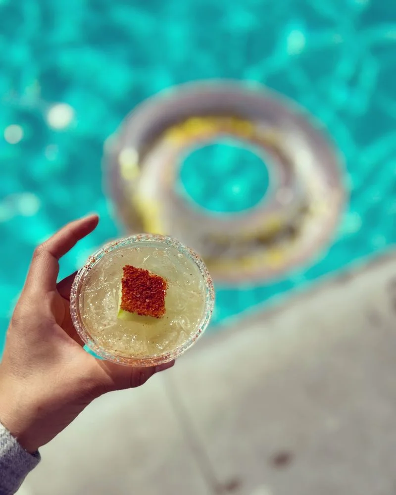 Two guests holding garnished cocktails over a swimming pool