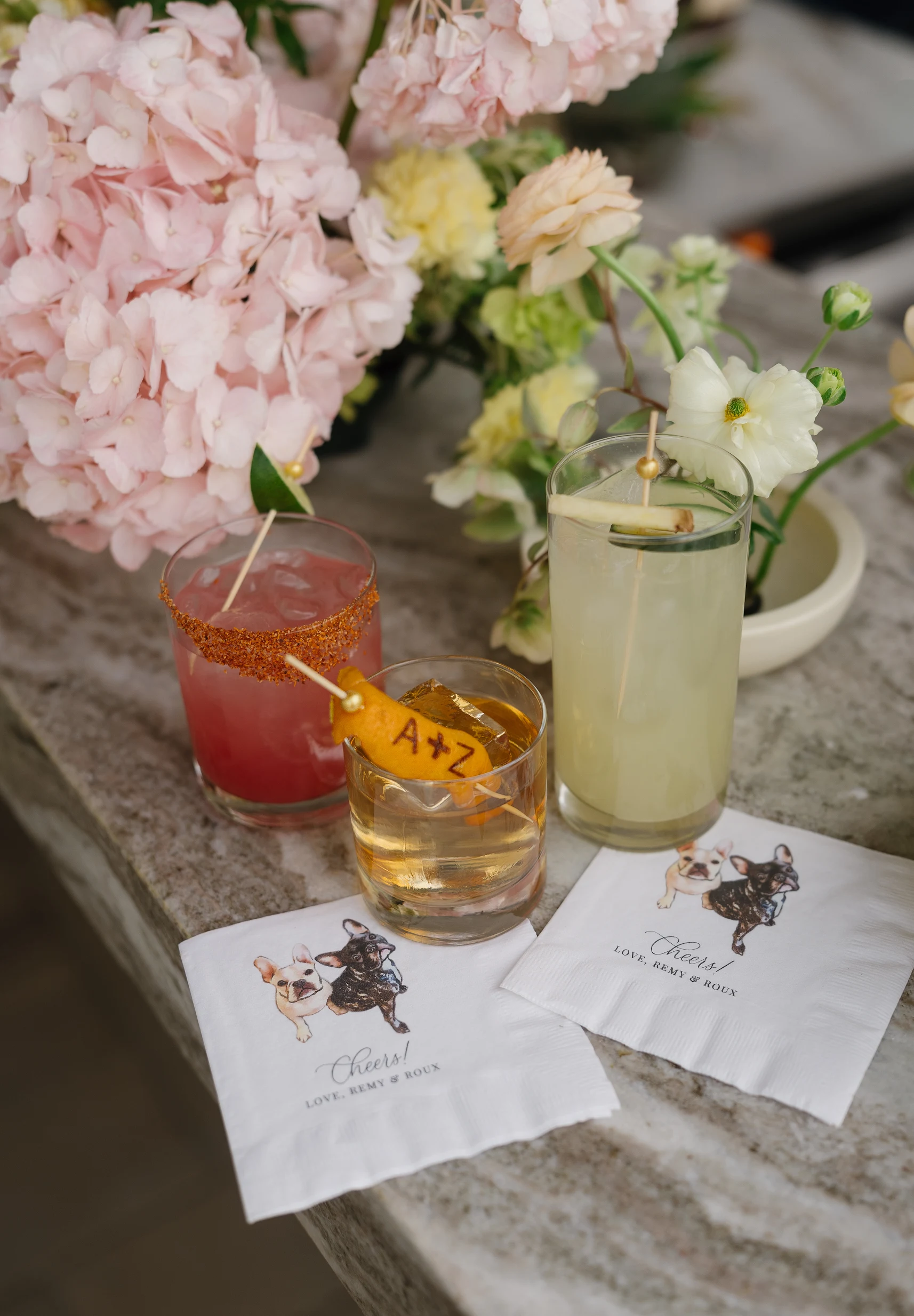 Three custom wedding cocktails beside pale pink flowers.