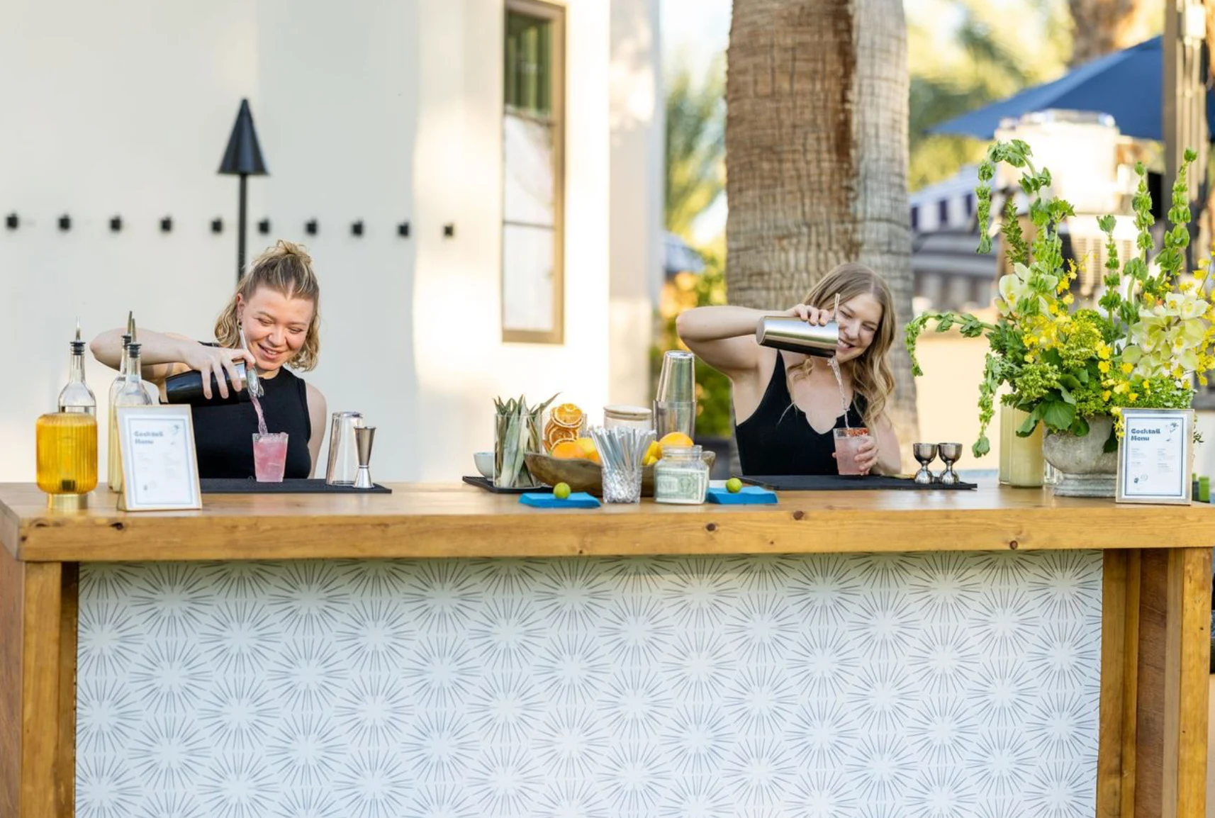 Two bartenders pouring pink cocktails together behind a polished outdoor mobile bar
