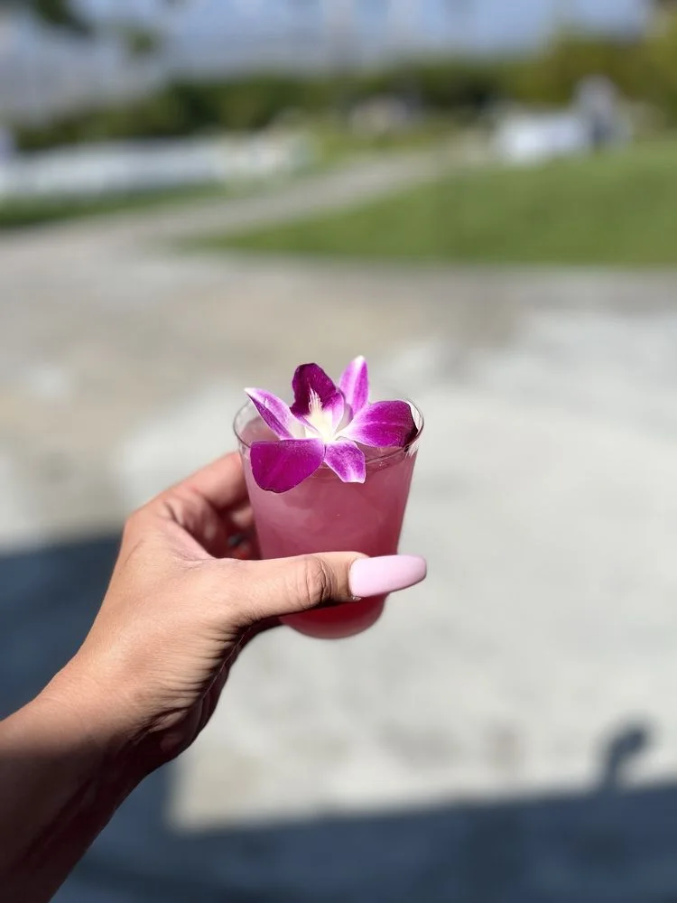 A hand holding a purple flower-topped cocktail beside a swimming pool.