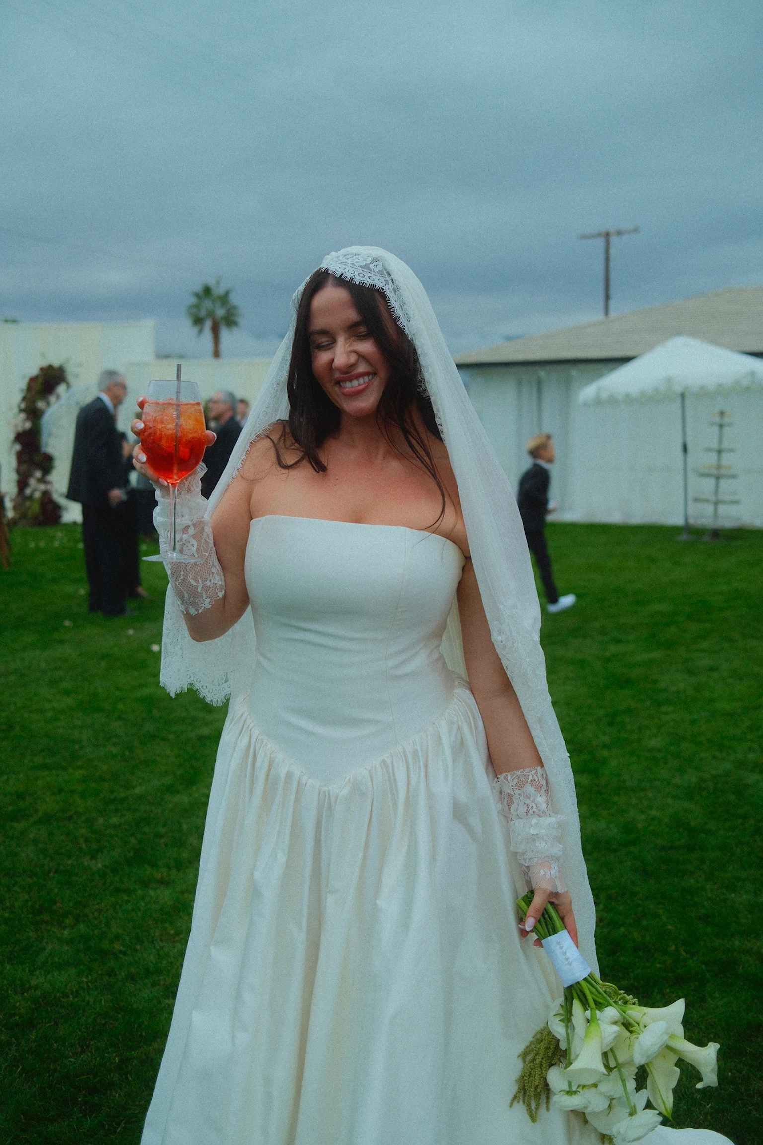 A bride smiling with a red signature cocktail at an outdoor wedding reception.